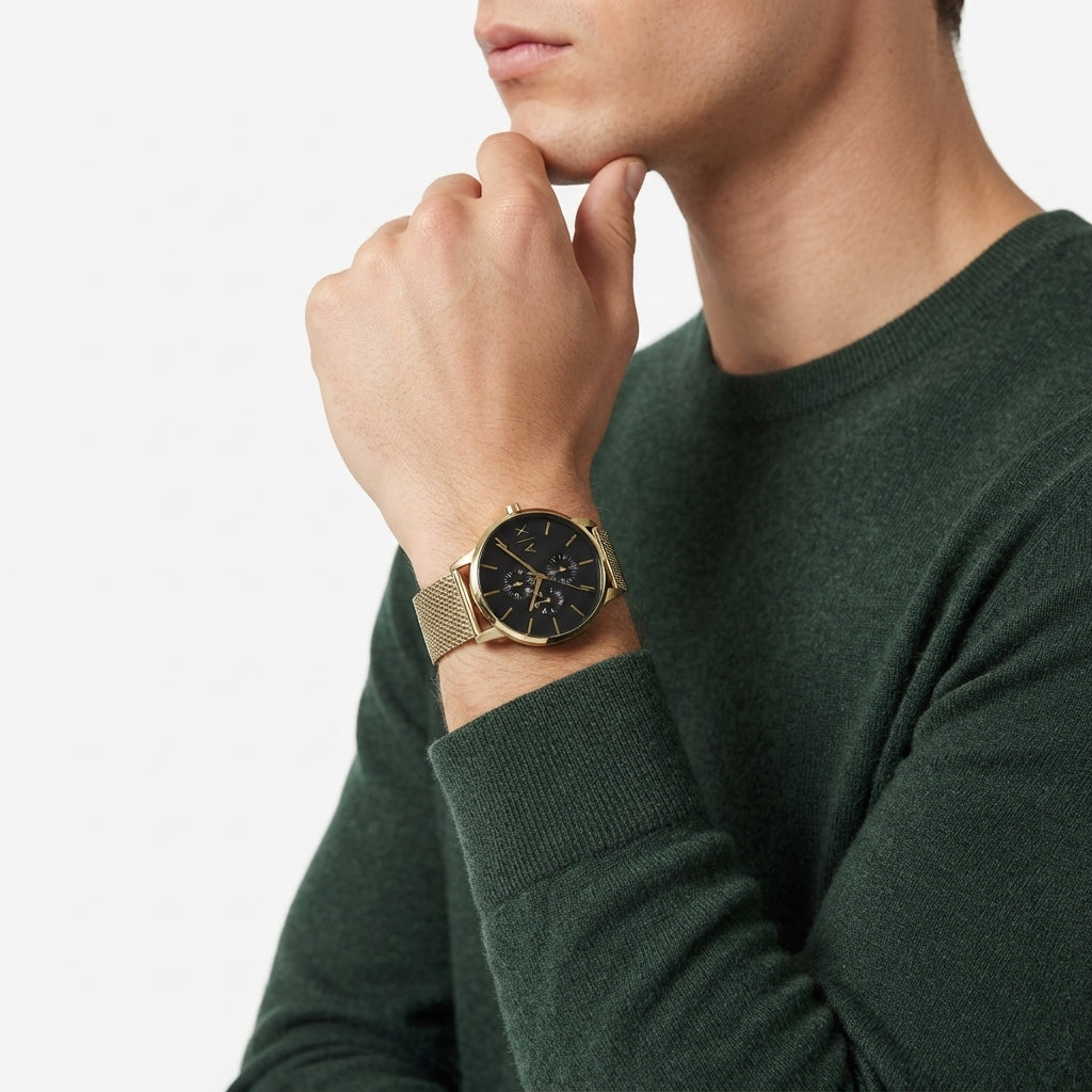 A man wearing a dark green knit sweater and a gold-tone mesh-strand watch with a black dial, shown in a close-up profile against a clean white background.