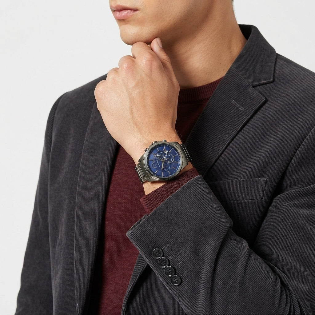 A close-up of a man wearing a silver-toned wristwatch with a blue dial and matching blue strap, styled with a dark gray blazer and burgundy sweater.