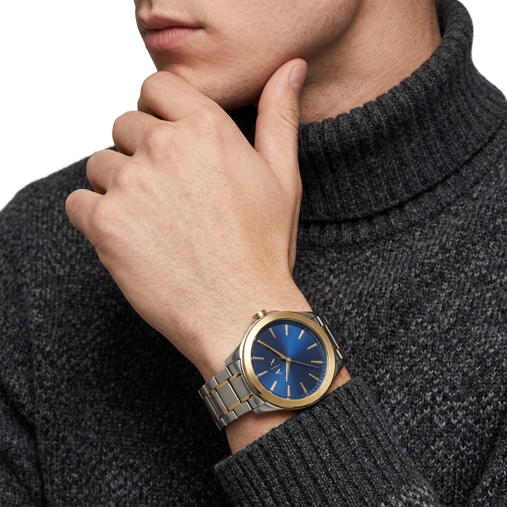 A men's wristwatch with a blue dial and silver-toned metal bracelet, shown worn on a person's wrist. The watch has a gold-tone bezel and is paired with a dark gray ribbed turtleneck sweater against a clean white background.