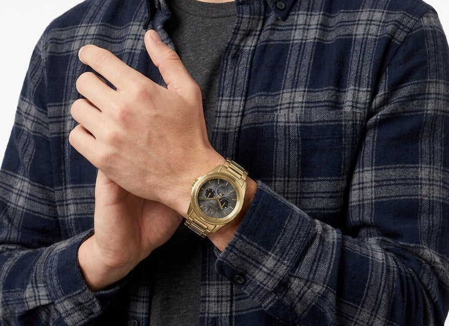 A man wearing a navy and gray plaid button-down shirt over a dark gray t-shirt, with a gold-tone wristwatch on his left wrist, against a plain white background.