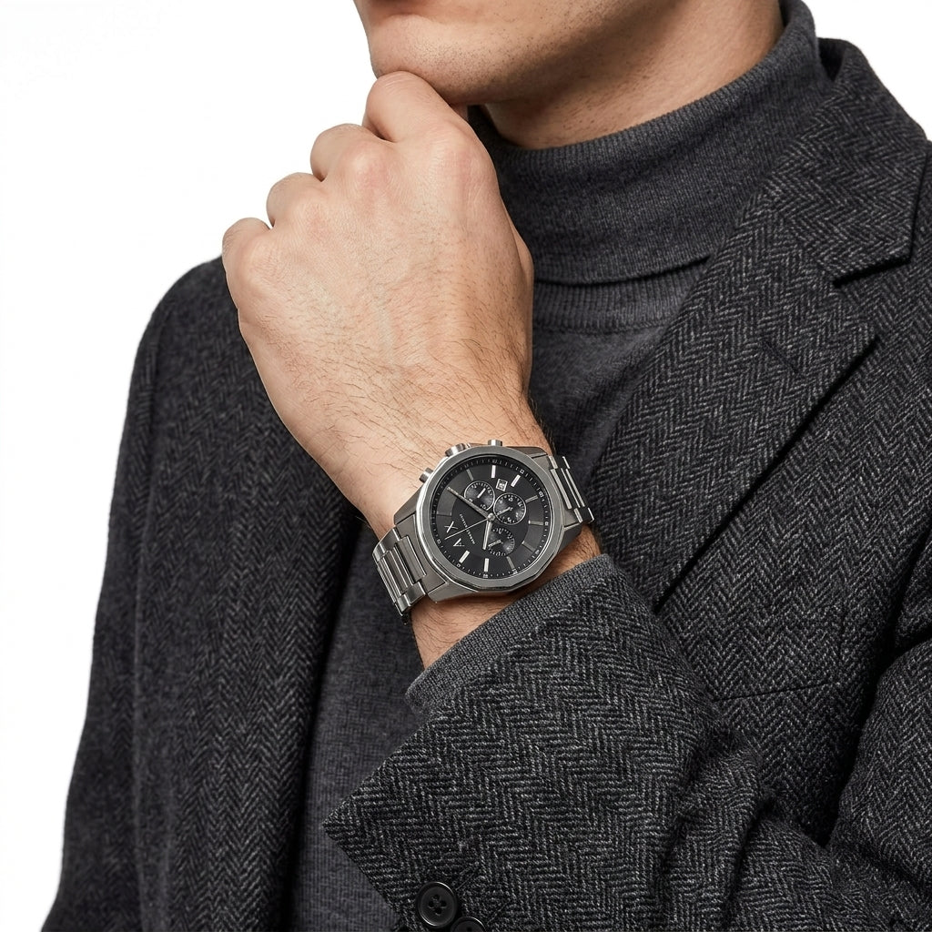 A close-up of a man wearing a silver-toned stainless steel watch with a black dial and a matching metal link bracelet, styled with a dark gray herringbone blazer and turtleneck sweater.