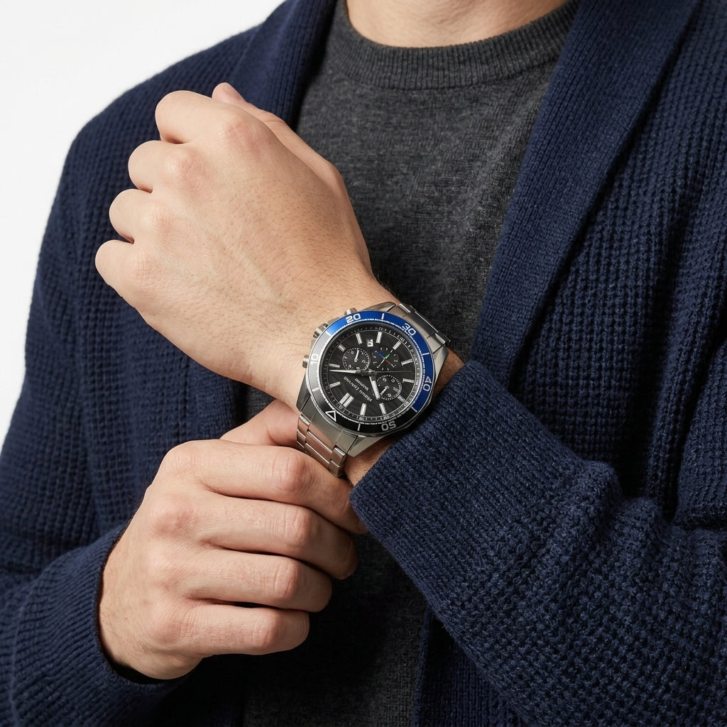 A close-up of a man wearing a silver stainless steel watch with a black dial and blue bezel, paired with a gray crewneck sweater and a navy blue knit cardigan.