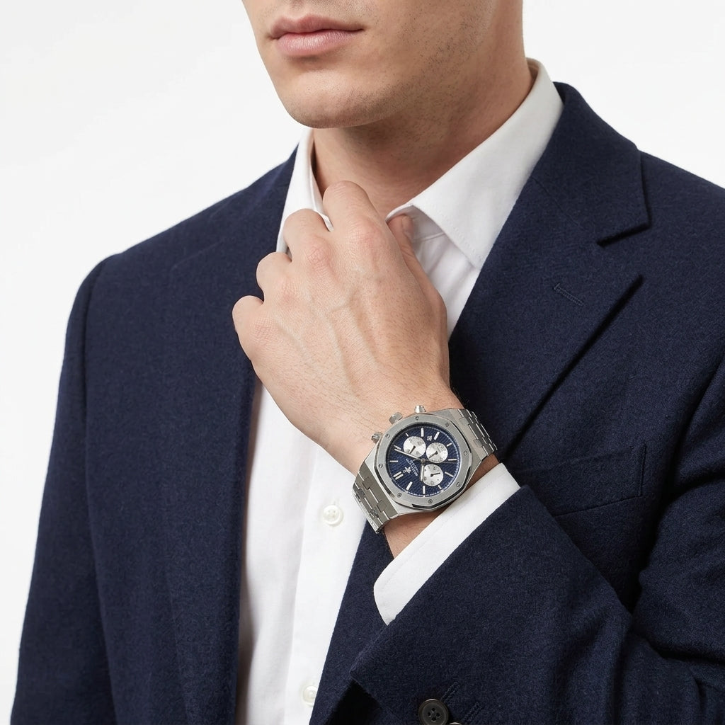 A silver-toned men's watch with a dark blue dial and three subdials, worn on the wrist of a man in a navy suit and white dress shirt.