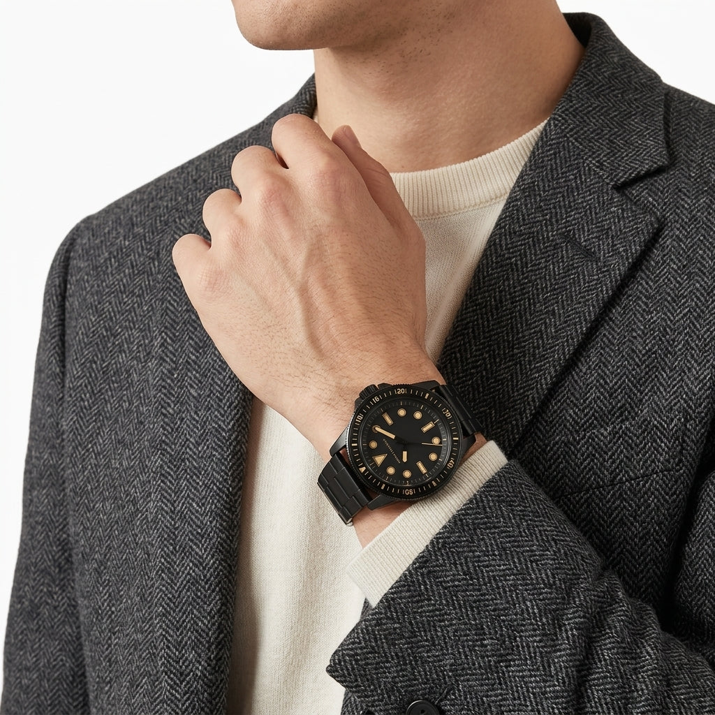 A close-up of a man wearing a black analog watch with a black strap and gold-tone hour markers on a dark gray herringbone blazer over a cream knit sweater.