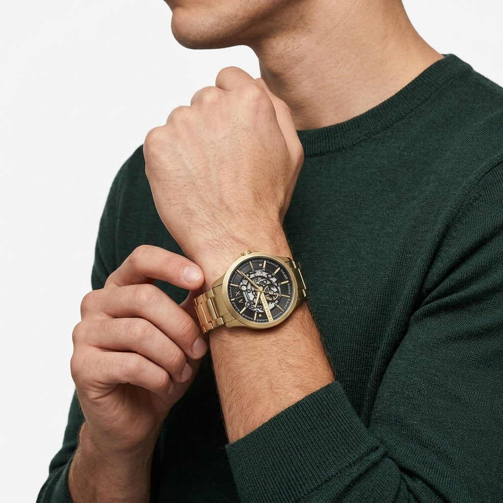 A close-up of a man wearing a gold-tone metal watch with a black dial and skeleton-style face, paired with a dark green crewneck sweater against a light gray background.